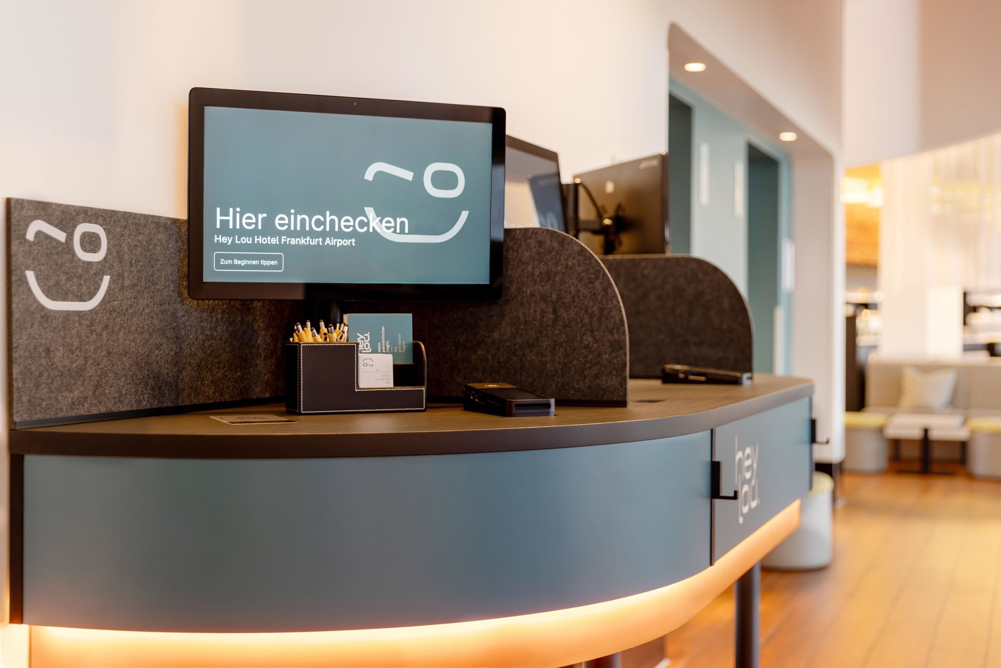 A reception desk with a digital screen displaying text in German, a small black box with pens, and a keycard holder.