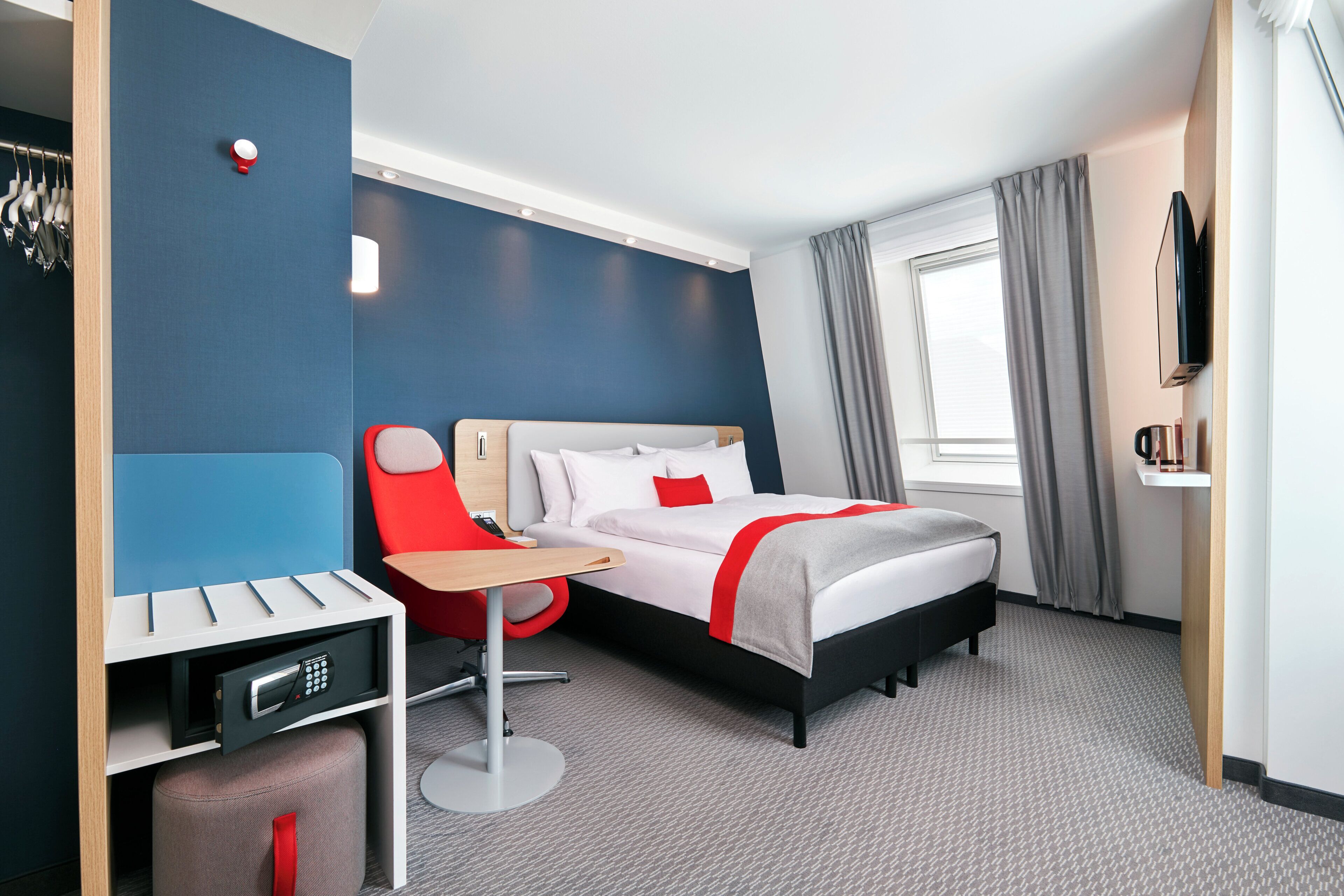 Una habitación de hotel con cama, escritorio, silla, televisión y ventana con cortinas.