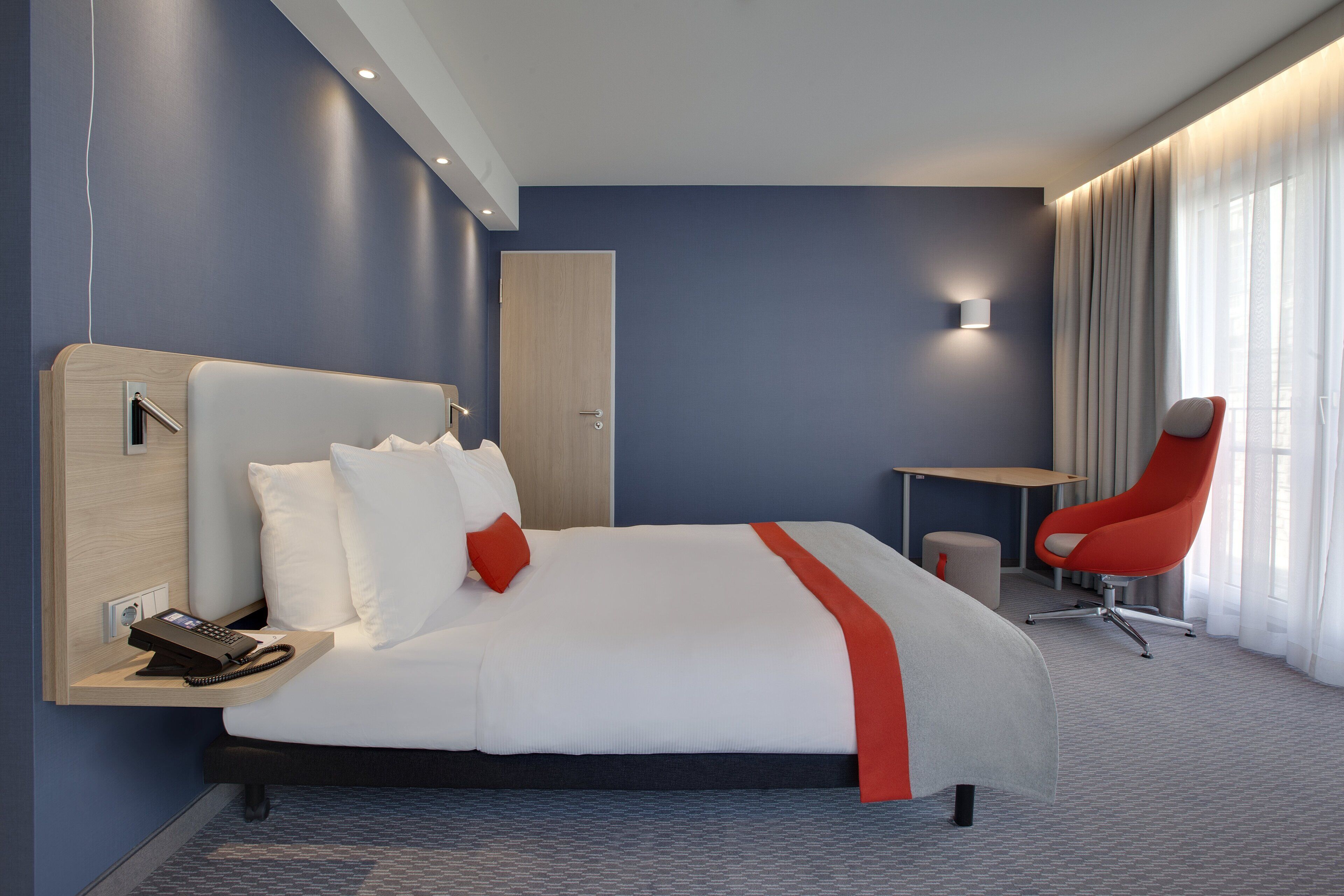 A modern hotel room with a large bed, a desk, a chair, and a phone on the nightstand.