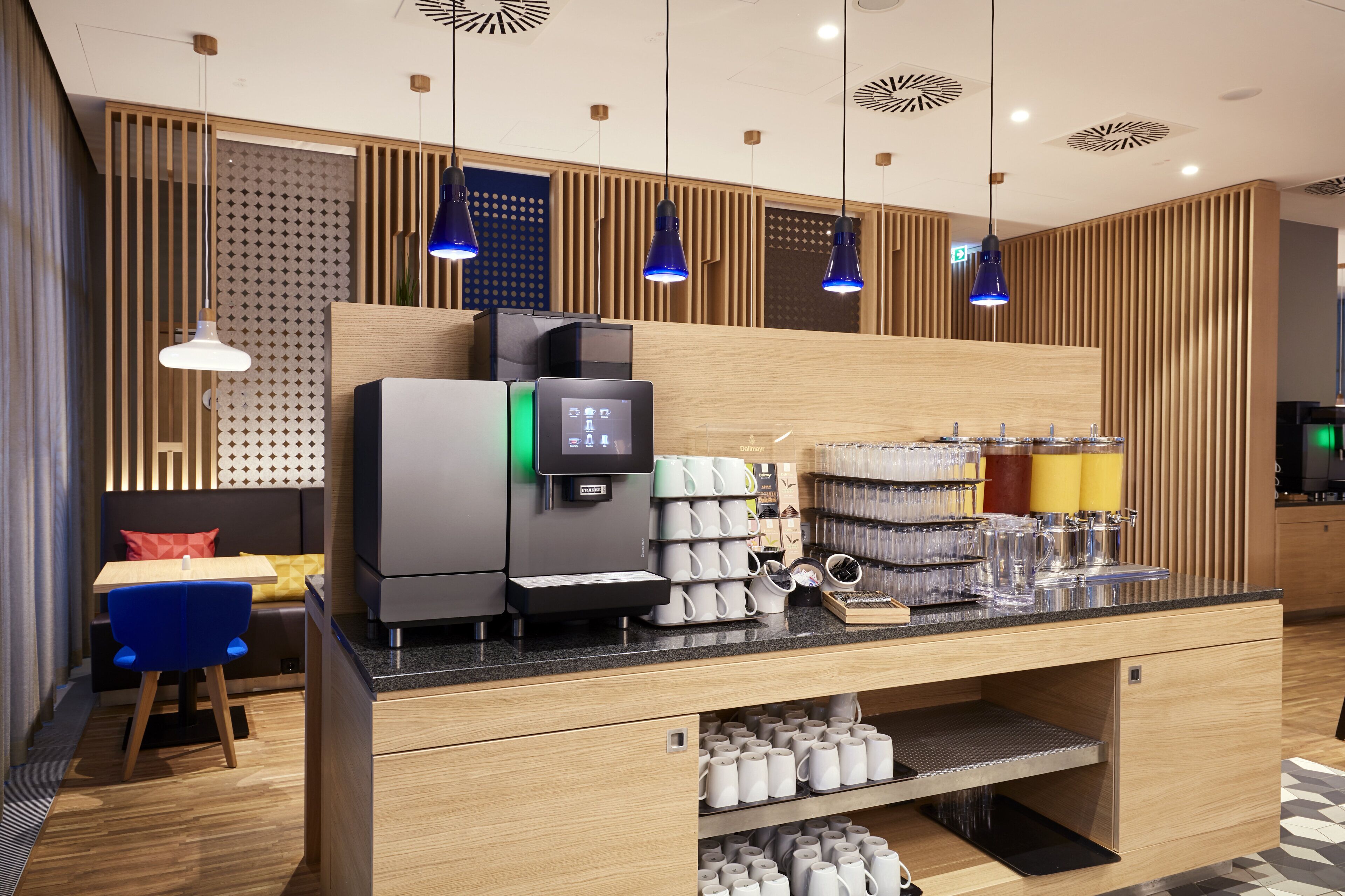Un espace café moderne comprenant une machine à café, des tasses et un coin salon avec une chaise bleue et un canapé.