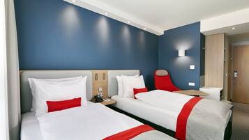 A hotel room with two beds, a red chair, a small table, and a wall-mounted light.