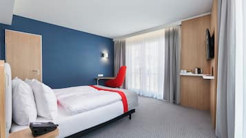 Una camera d'albergo con un letto, una poltrona rossa, un televisore fissato al muro e una scrivania con un telefono.