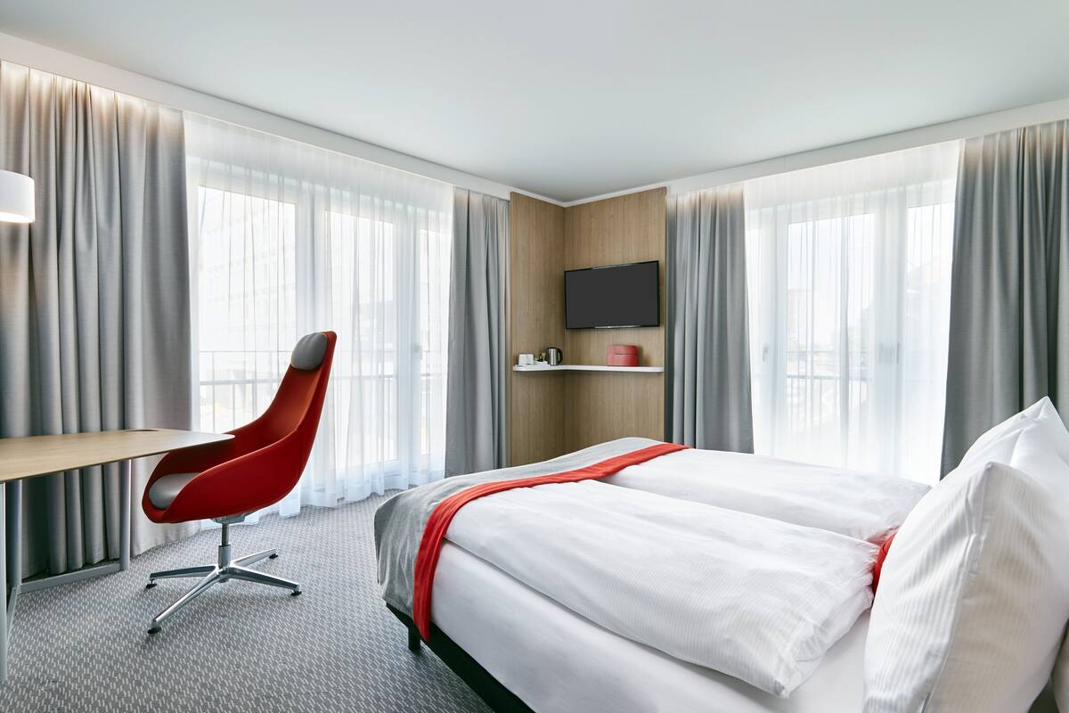 A hotel room with a bed, a desk, a chair, a TV, and large windows with curtains.