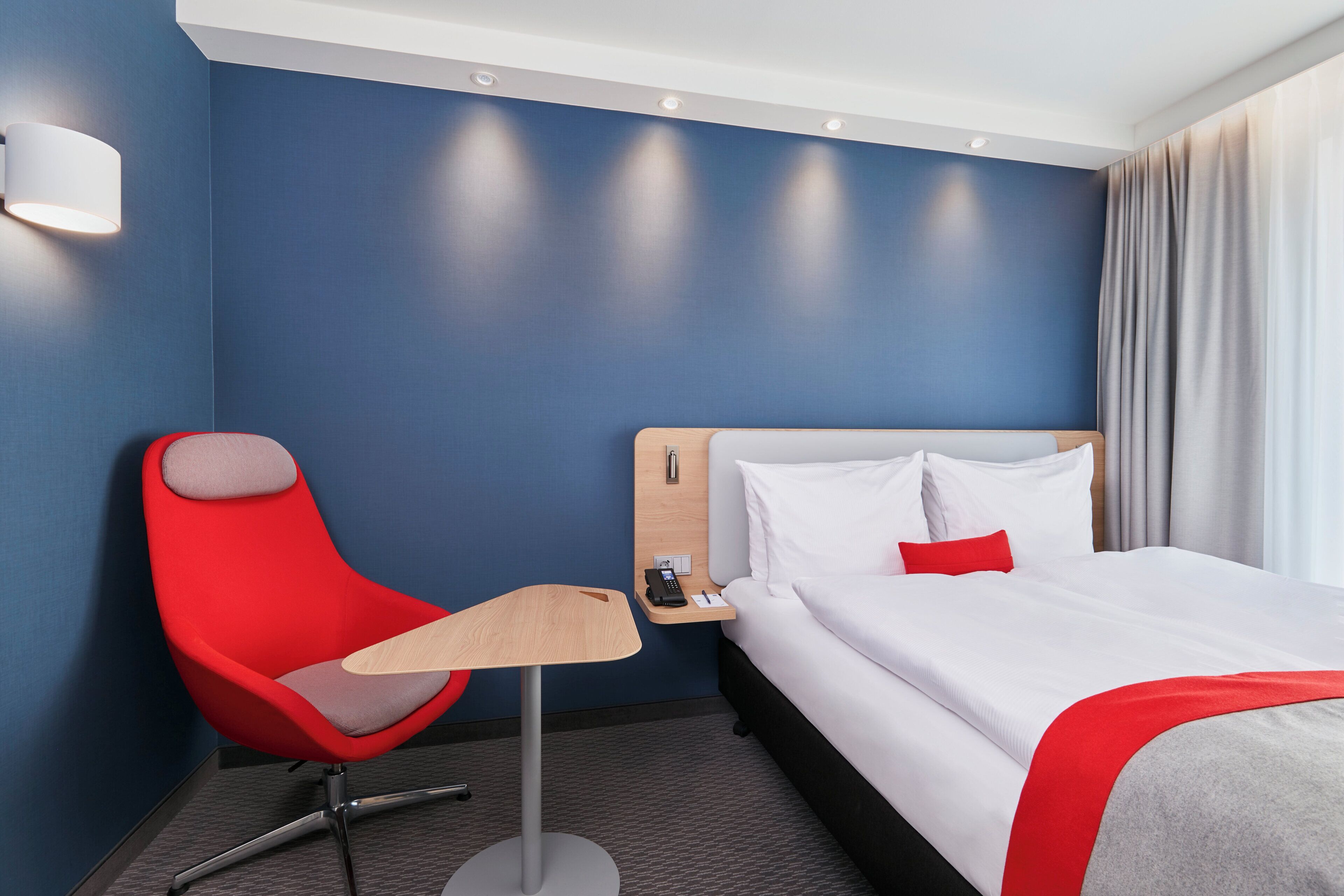 Quarto de hotel com uma cama de solteiro, uma cadeira vermelha, uma mesa pequena e uma mesinha de cabeceira com telefone e abajur.