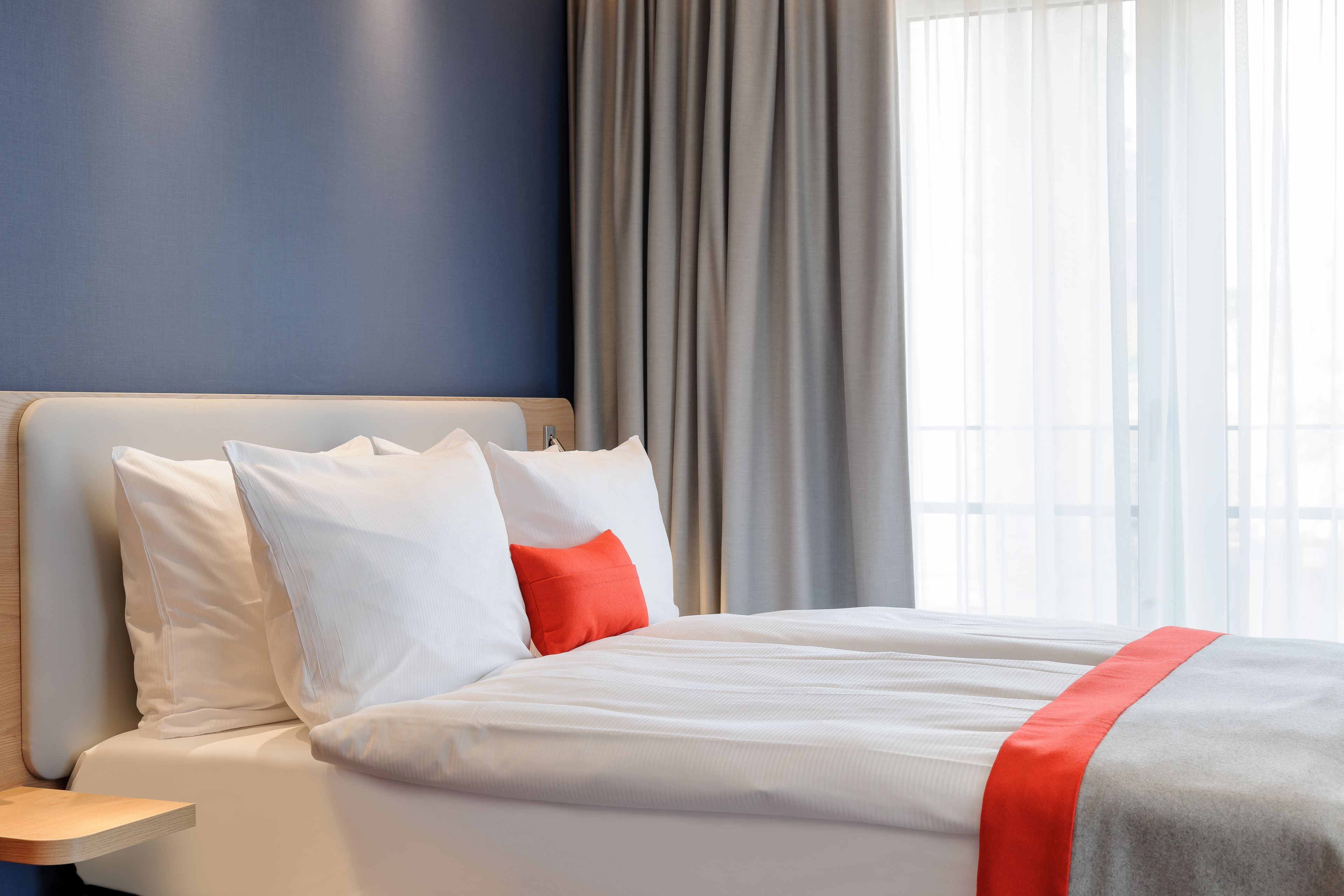 Ett hotellrum med en säng, vita sängkläder, en röd prydnadskudde och utsikt över omgivningarna genom stora fönster.