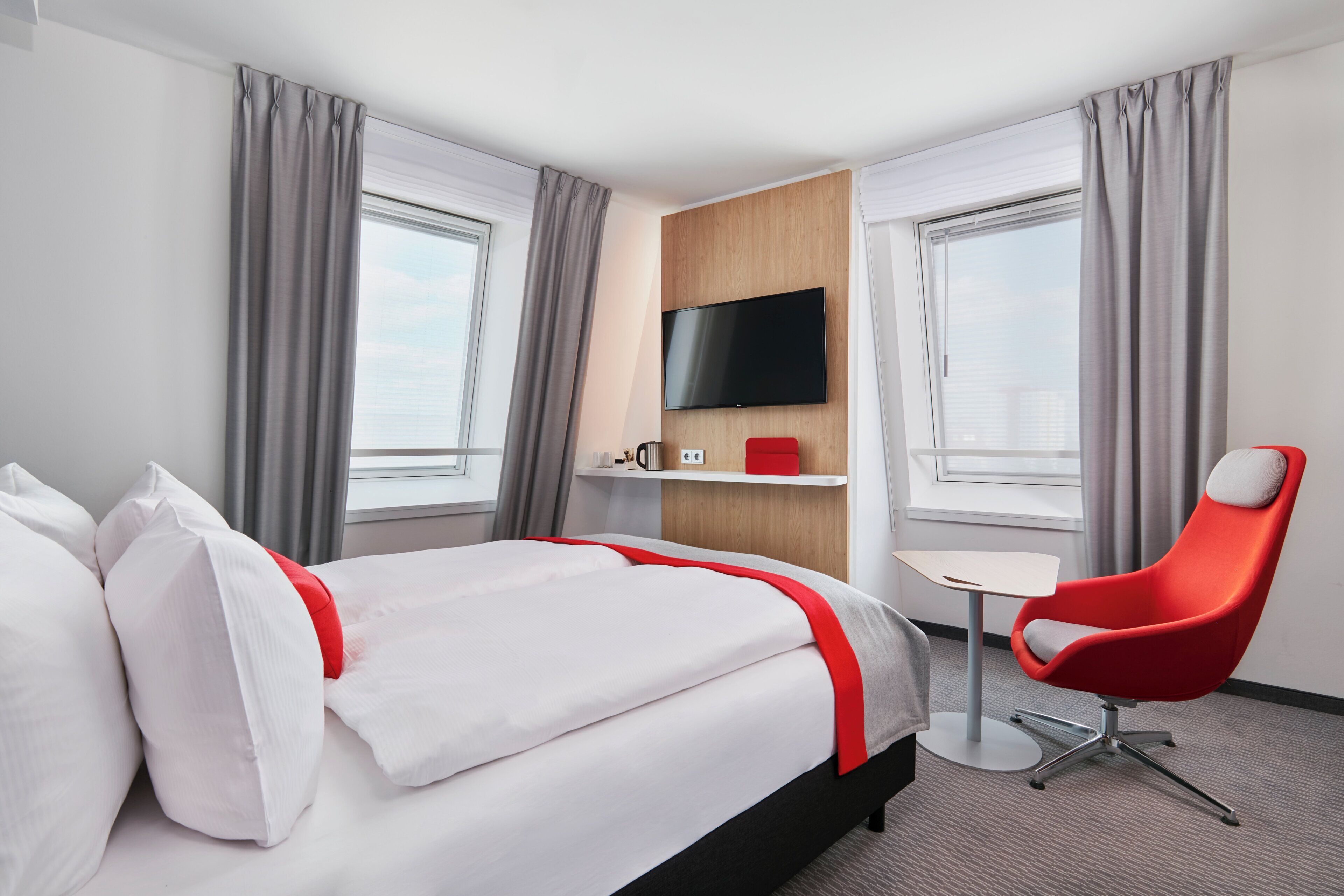 A hotel room with a bed, a red chair, a small table, a TV, and a window.