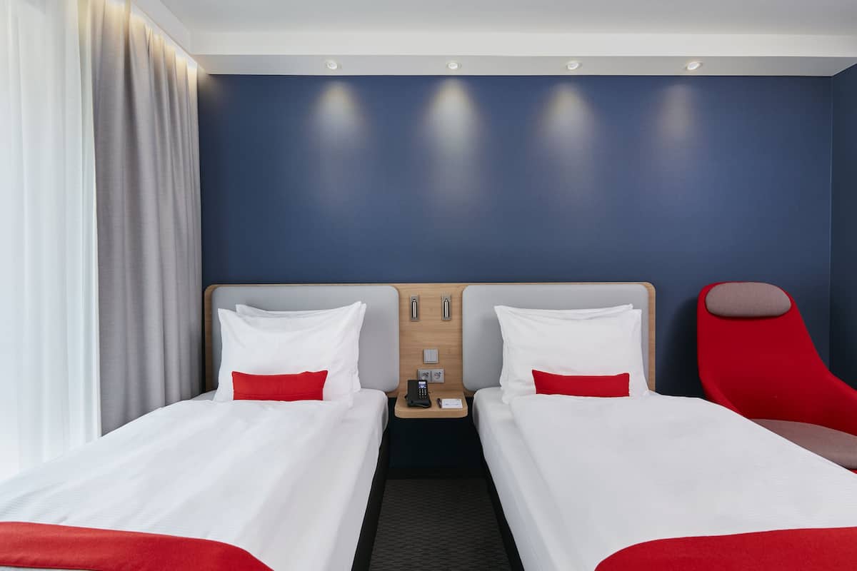 A hotel room with two beds, a red chair, a nightstand with a phone, and a wall with a blue accent.
