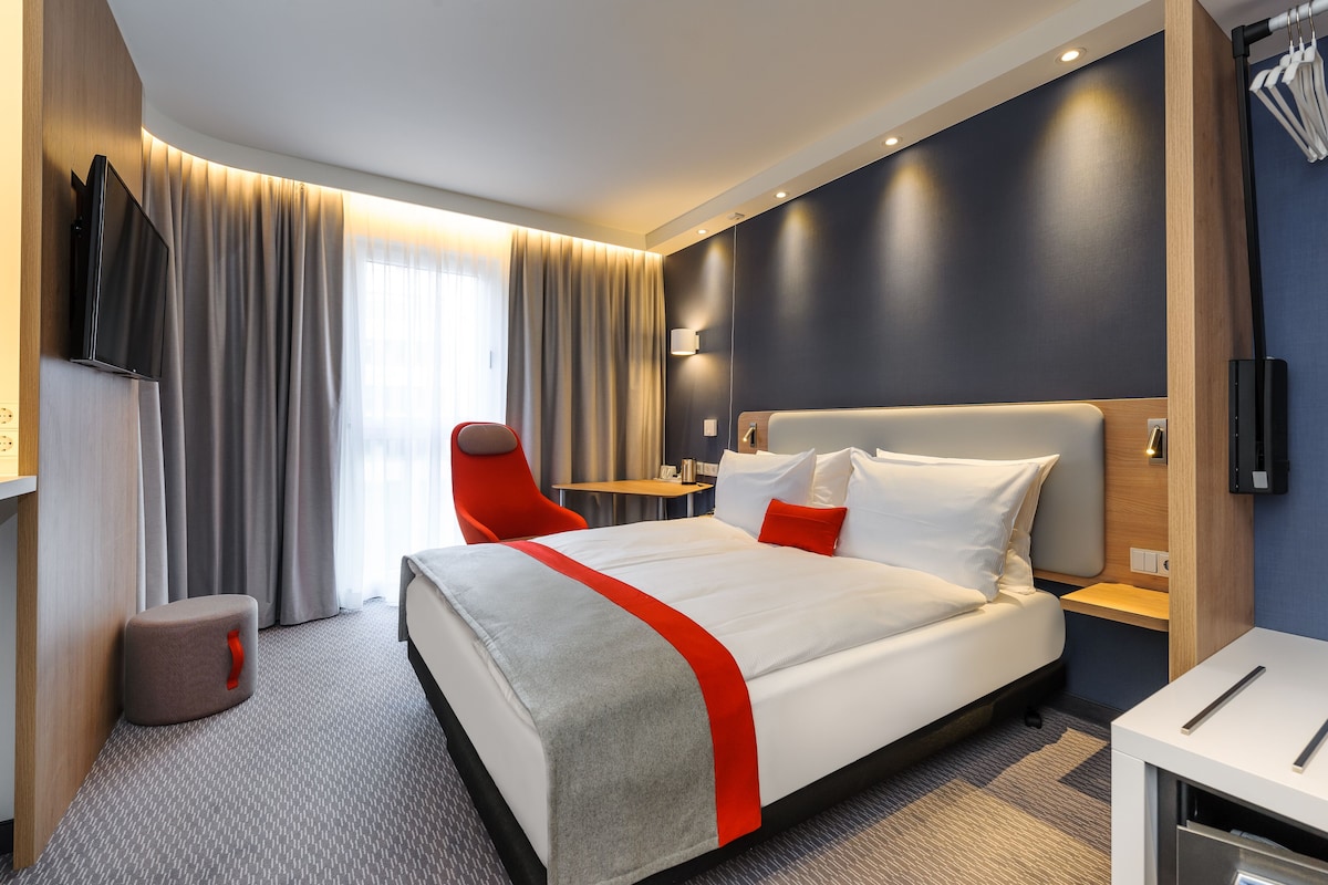 Ein modernes Hotelzimmer mit einem großen Bett, einem roten Akzentstuhl, einem Flachbildfernseher und einem Nachttisch.
