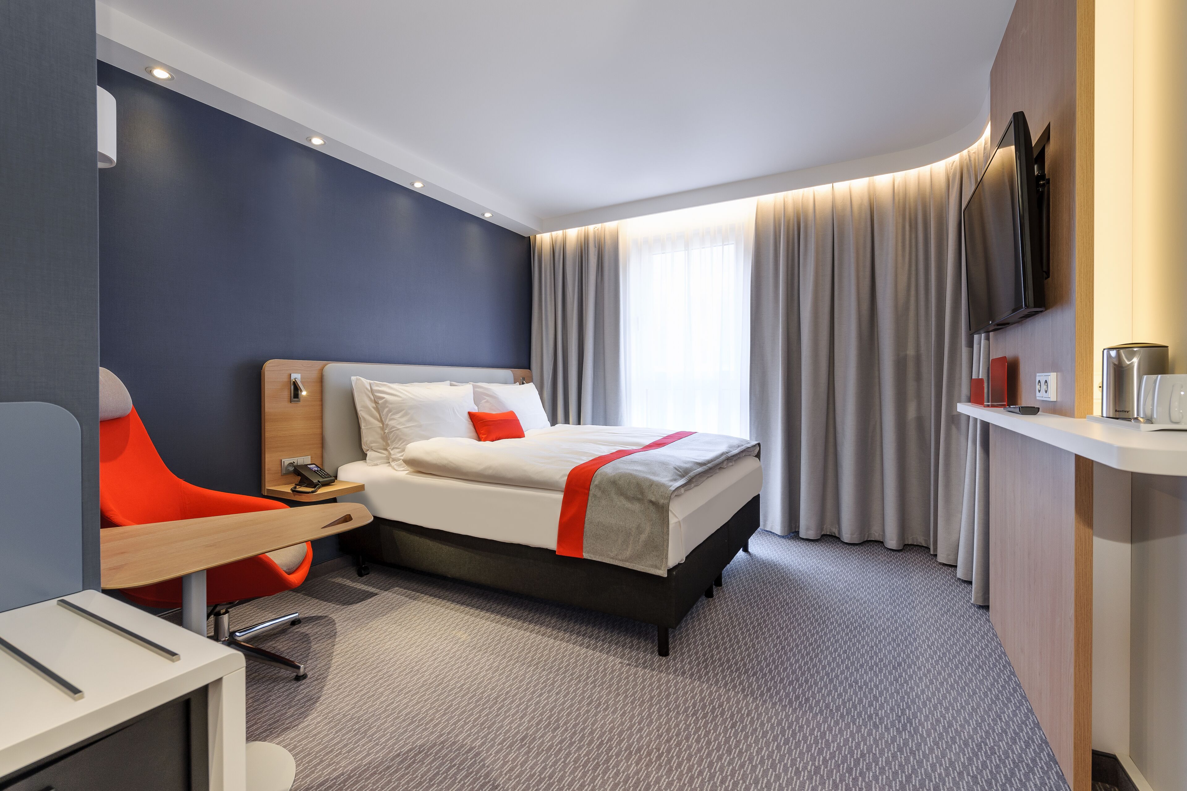 A modern hotel room with a bed, a desk, a chair, a TV, and a window with curtains.