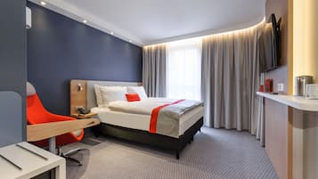 A modern hotel room with a bed, a desk, a chair, a TV, and a window with curtains.