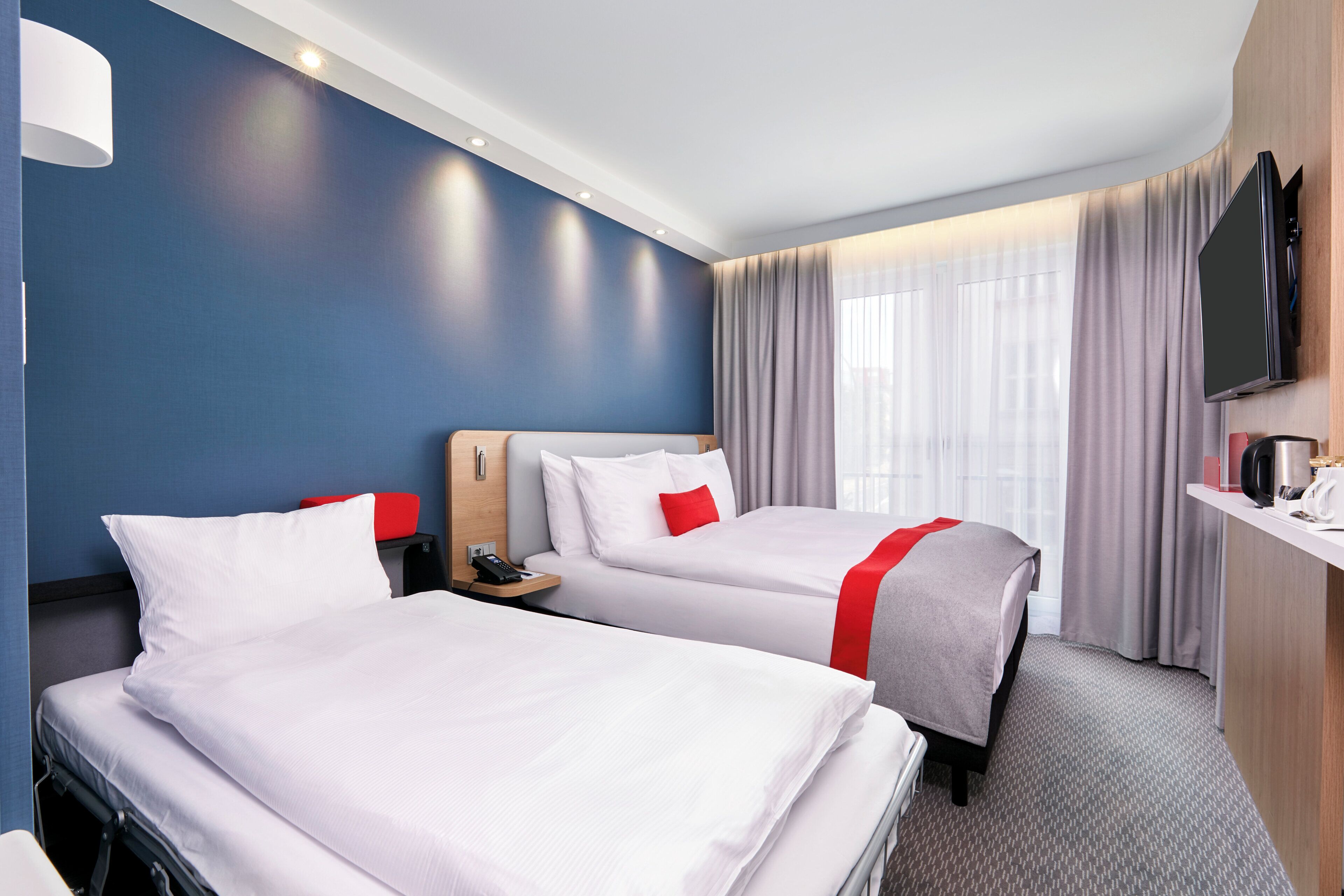 A hotel room with two beds, a desk, a TV, and a window with curtains.