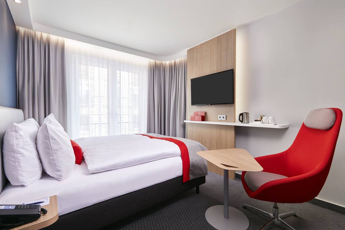 A modern hotel room with a bed, a red chair, a small table, a TV, and a coffee machine.