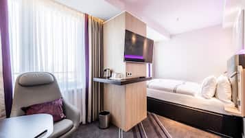 Una habitación de hotel moderna con una cama, un televisor montado en un panel de madera, un escritorio pequeño con un hervidor, una silla y un bote de basura.