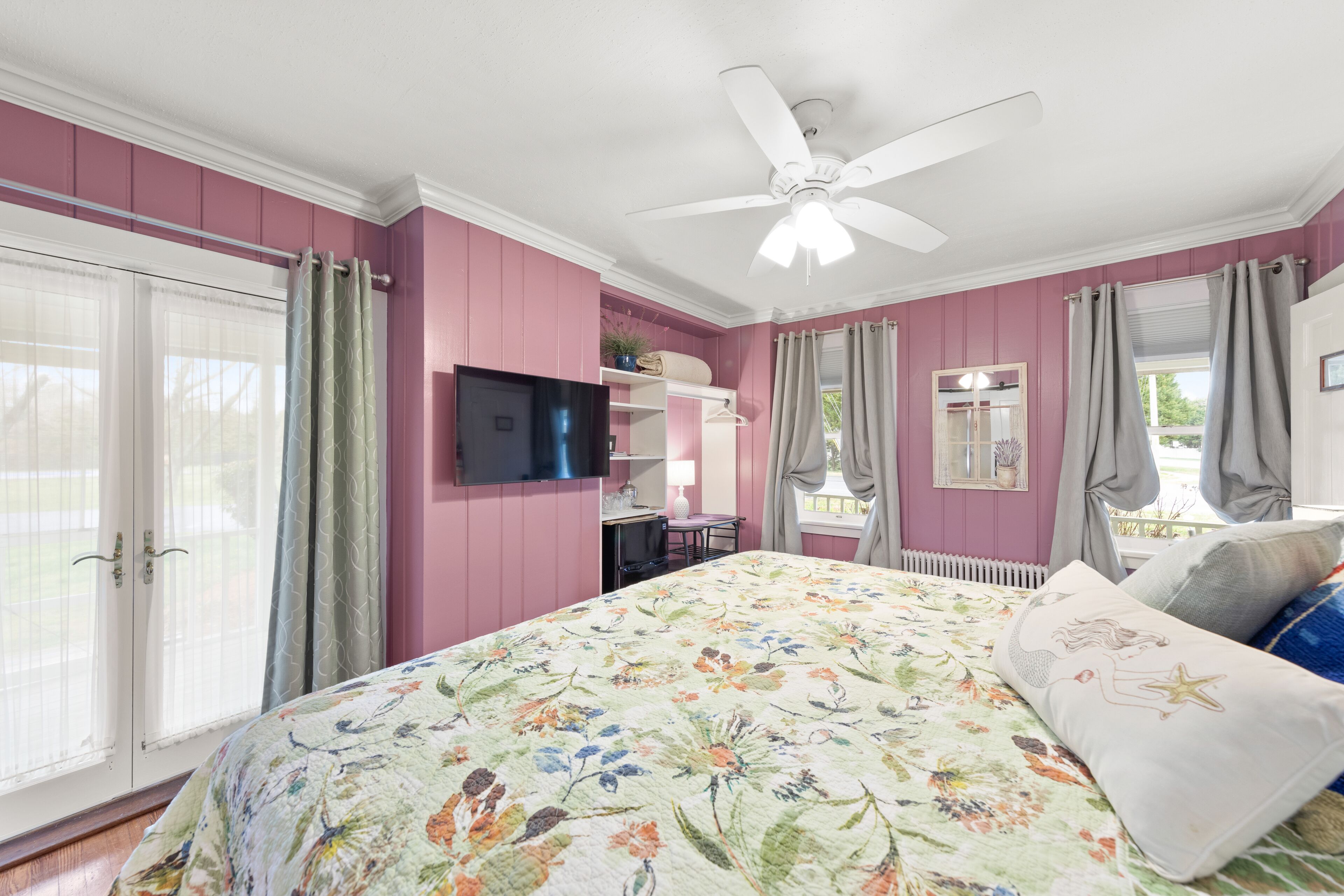 A bedroom with a floral bedspread, a ceiling fan, a TV, and a window with curtains.