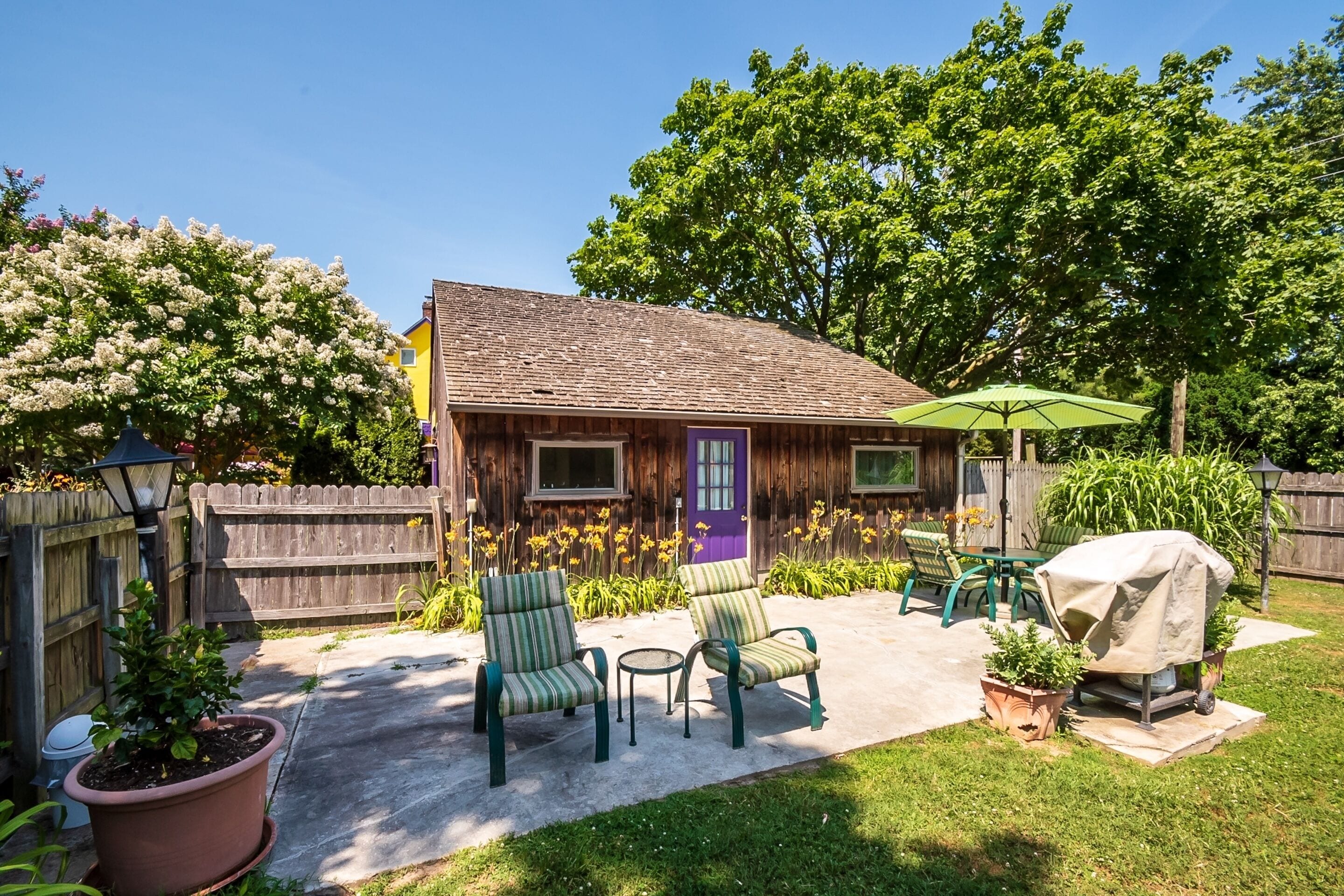 Ein Holzhaus mit einer violetten Tür, einer Terrasse mit grünen Stühlen, einem Grill und Kübelpflanzen.