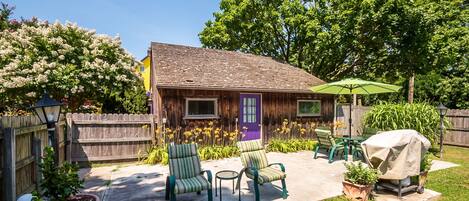 Ein Holzhaus mit einer violetten Tür, einer Terrasse mit grünen Stühlen, einem Grill und Kübelpflanzen.