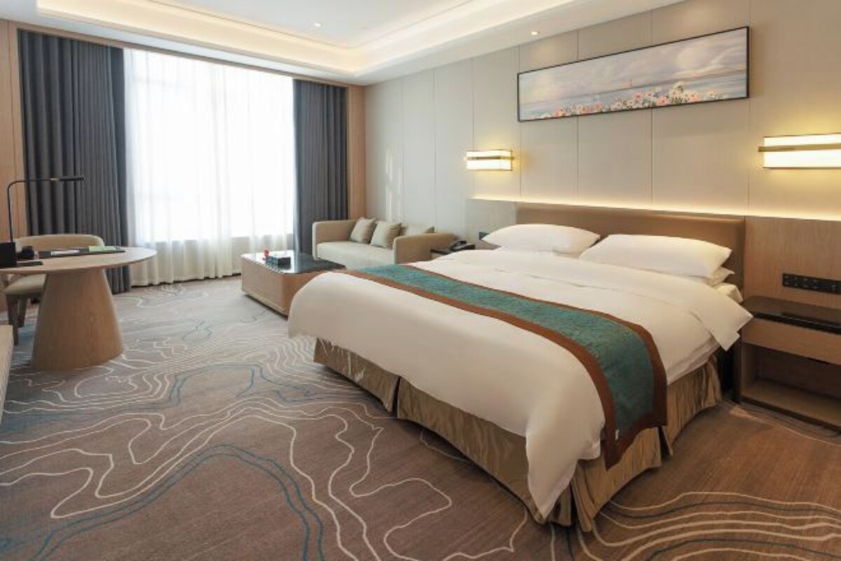 A hotel room with a large bed, a desk, a sofa, and a view of the outdoors through the window.