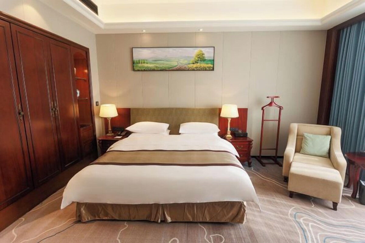 Habitación de hotel con una cama grande, dos mesitas de noche con lámparas, un armario de madera, una silla y un cuadro en la pared.