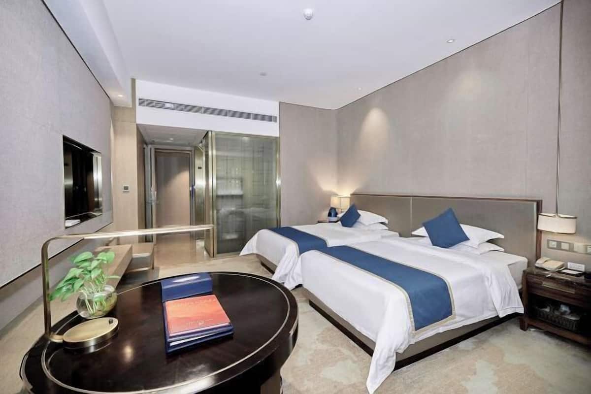 A hotel room with two beds, a desk with a chair, a TV, and a glass door leading to a shower area.