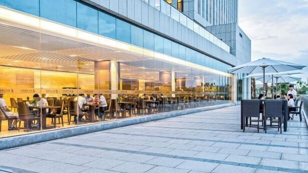 An outdoor dining area with tables and chairs, a glass facade restaurant, and a modern building in the background. - Fontaine Hotel (Yingtan)