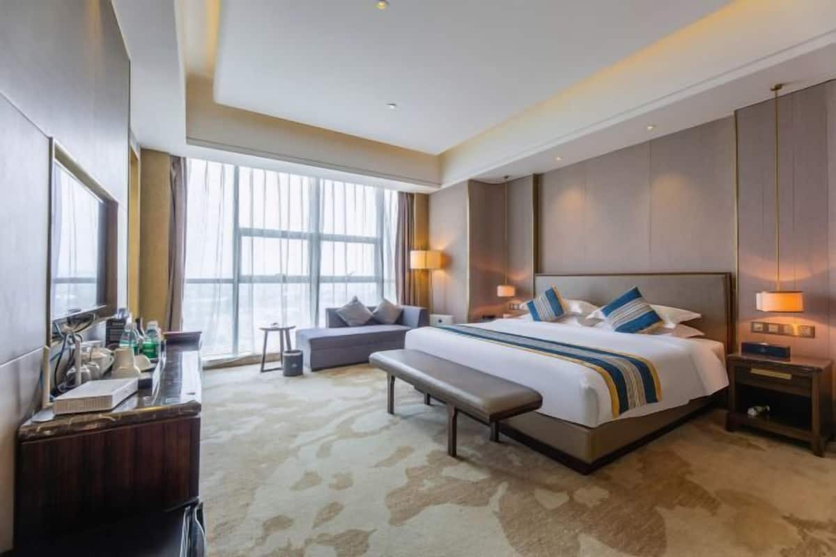 A hotel room with a large bed, a bench, a desk, and a window with curtains.