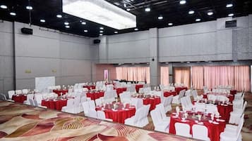 Un amplio salón de banquetes con filas de mesas y sillas, una alfombra estampada y un techo alto con luminarias.