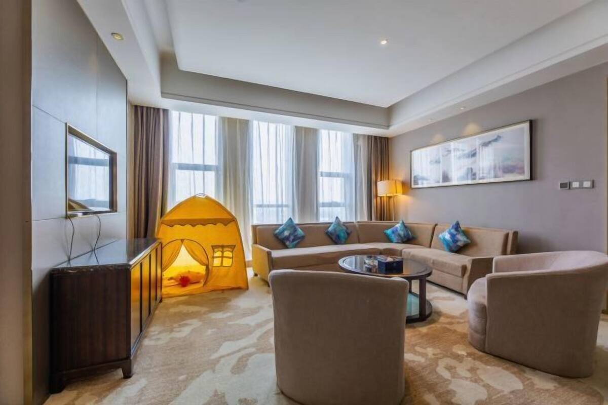 A modern hotel room with a sofa set, a small tent-like structure, a TV, and a large window with curtains.