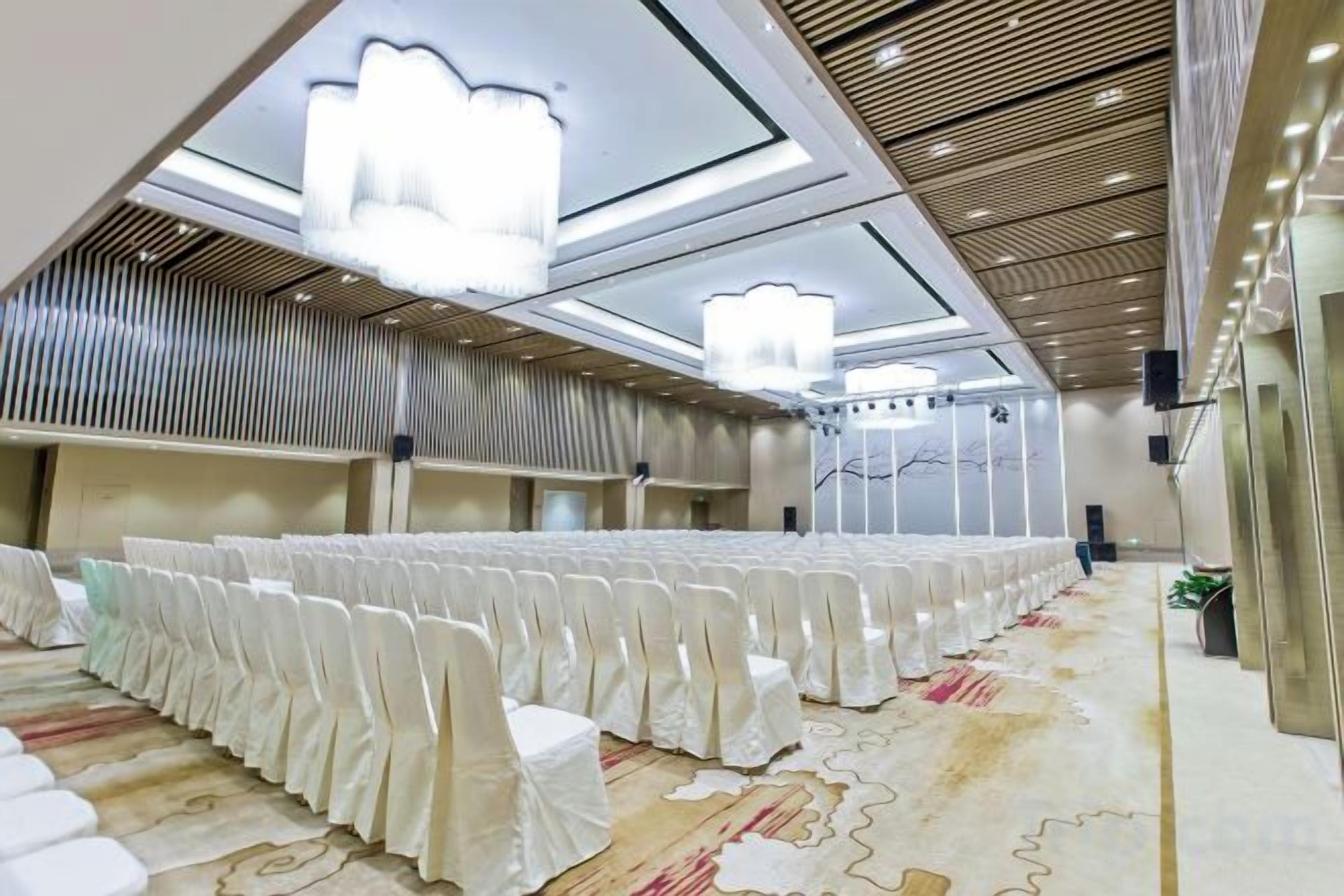A banquet hall with rows of white-covered chairs, a patterned carpet, and modern chandeliers.