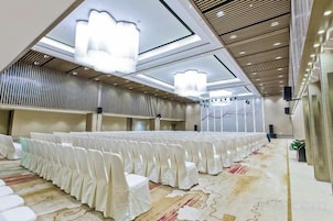 Un salón de banquetes con filas de sillas cubiertas de blanco, una alfombra estampada y candelabros modernos.