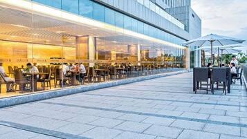 Un área de comedor al aire libre con mesas y sillas, fachada de edificio moderna y cielo despejado.
