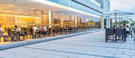 An outdoor dining area with tables and chairs, a modern building facade, and a clear sky.