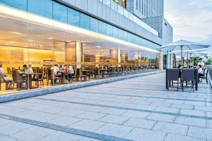 Un área de comedor al aire libre con mesas y sillas, fachada de edificio moderna y cielo despejado.