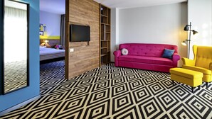Ein Hotelzimmer mit Bett, einem an einer Holzplatte montierten Fernseher, einem rosafarbenen Sofa, einem gelben Sessel und einem gemusterten Boden.