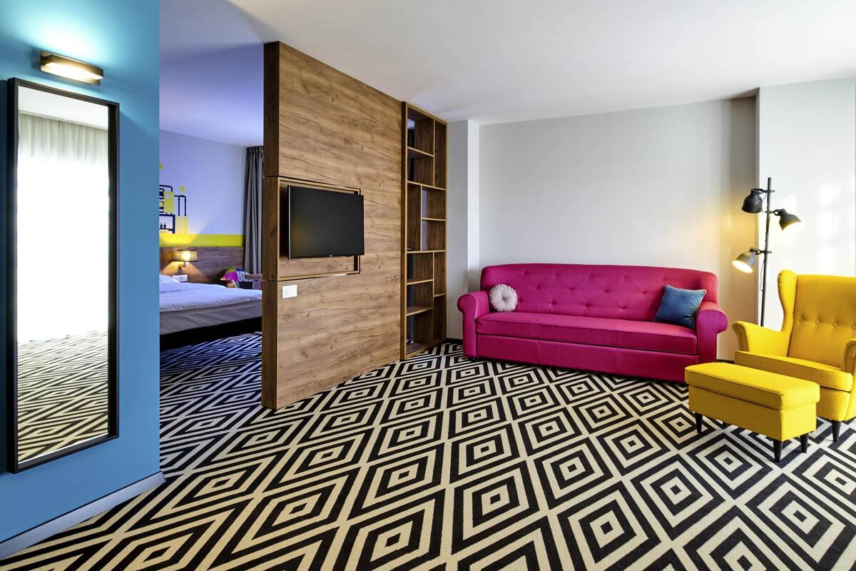 A hotel room with a bed, a TV mounted on a wooden panel, a pink sofa, a yellow armchair, and a patterned floor.