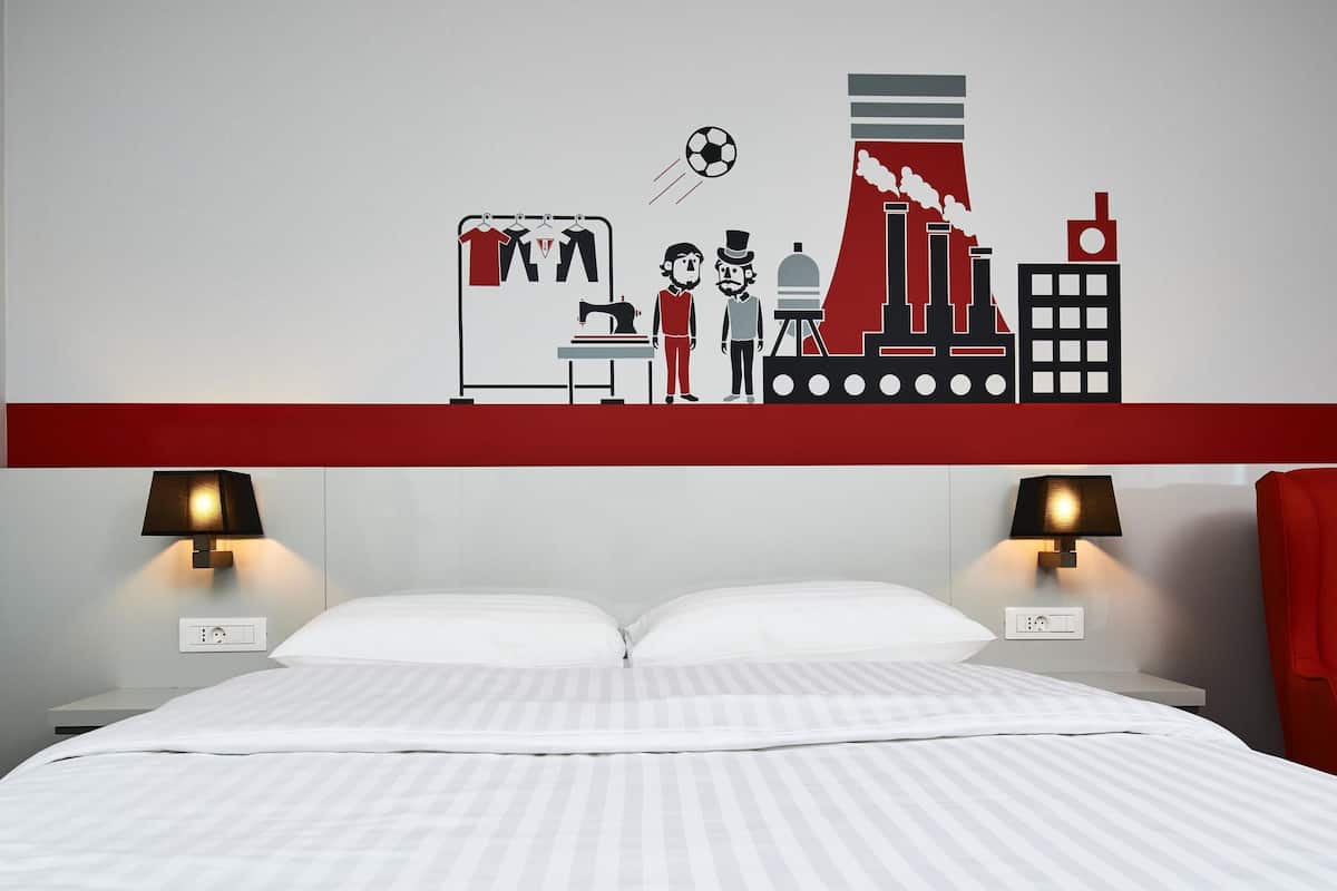 A hotel room with a bed, two bedside lamps, a red-striped wall, and a wall mural depicting industrial scenes.