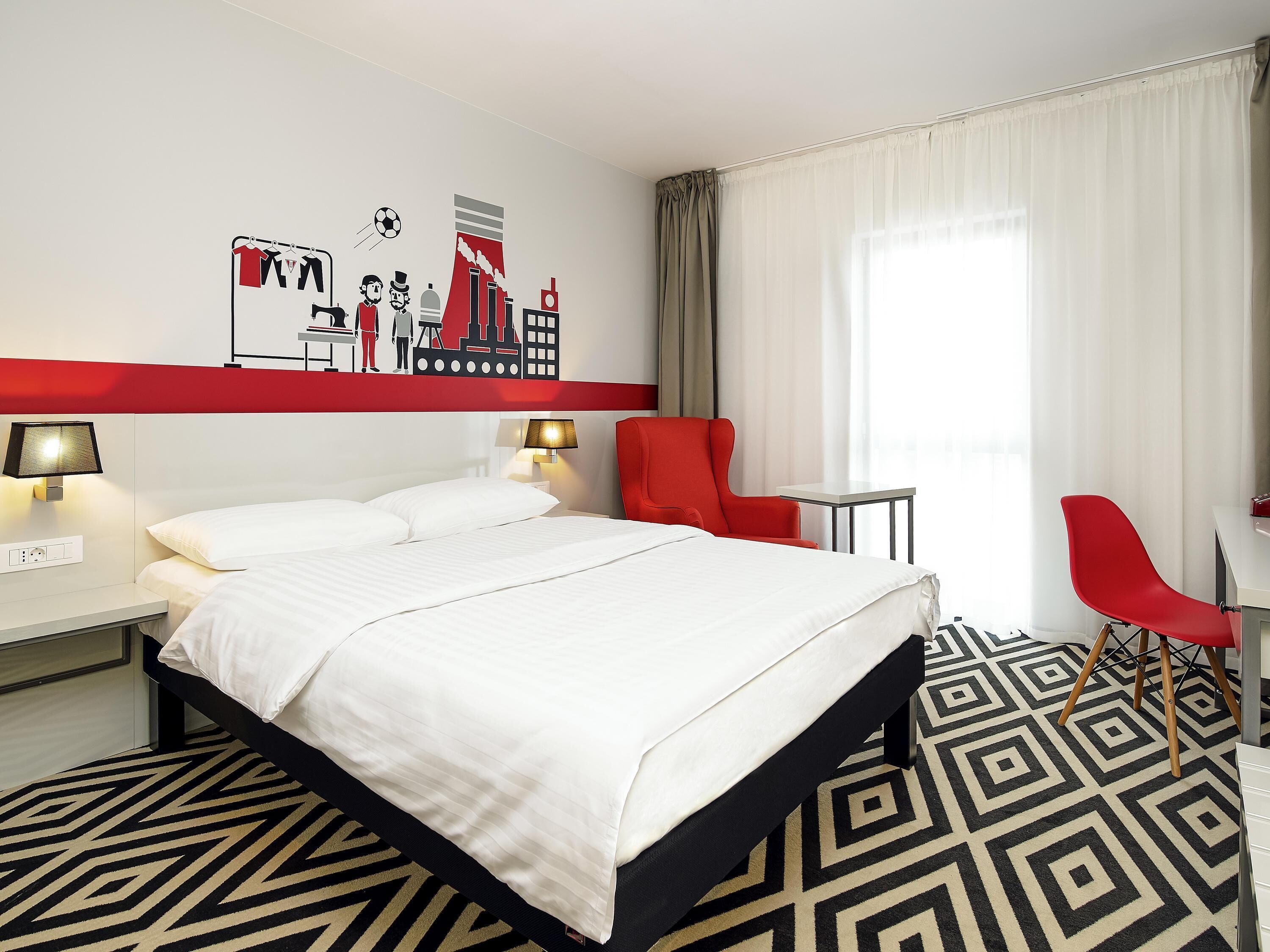 Una habitació d'hotel amb un llit, dues làmpades de nit, una cadira vermella, una taula petita i un mural a la paret.