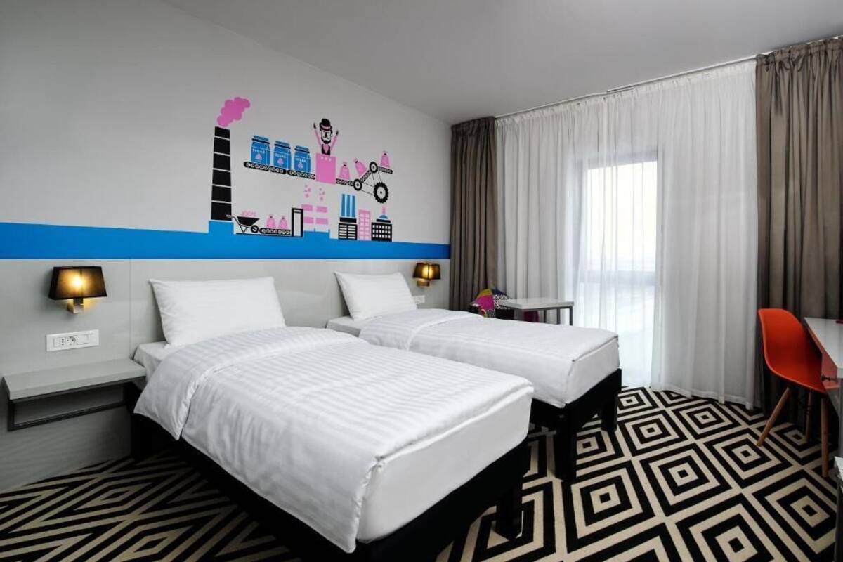 A hotel room with two beds, a desk, a chair, a wall with a cityscape mural, and a patterned floor.