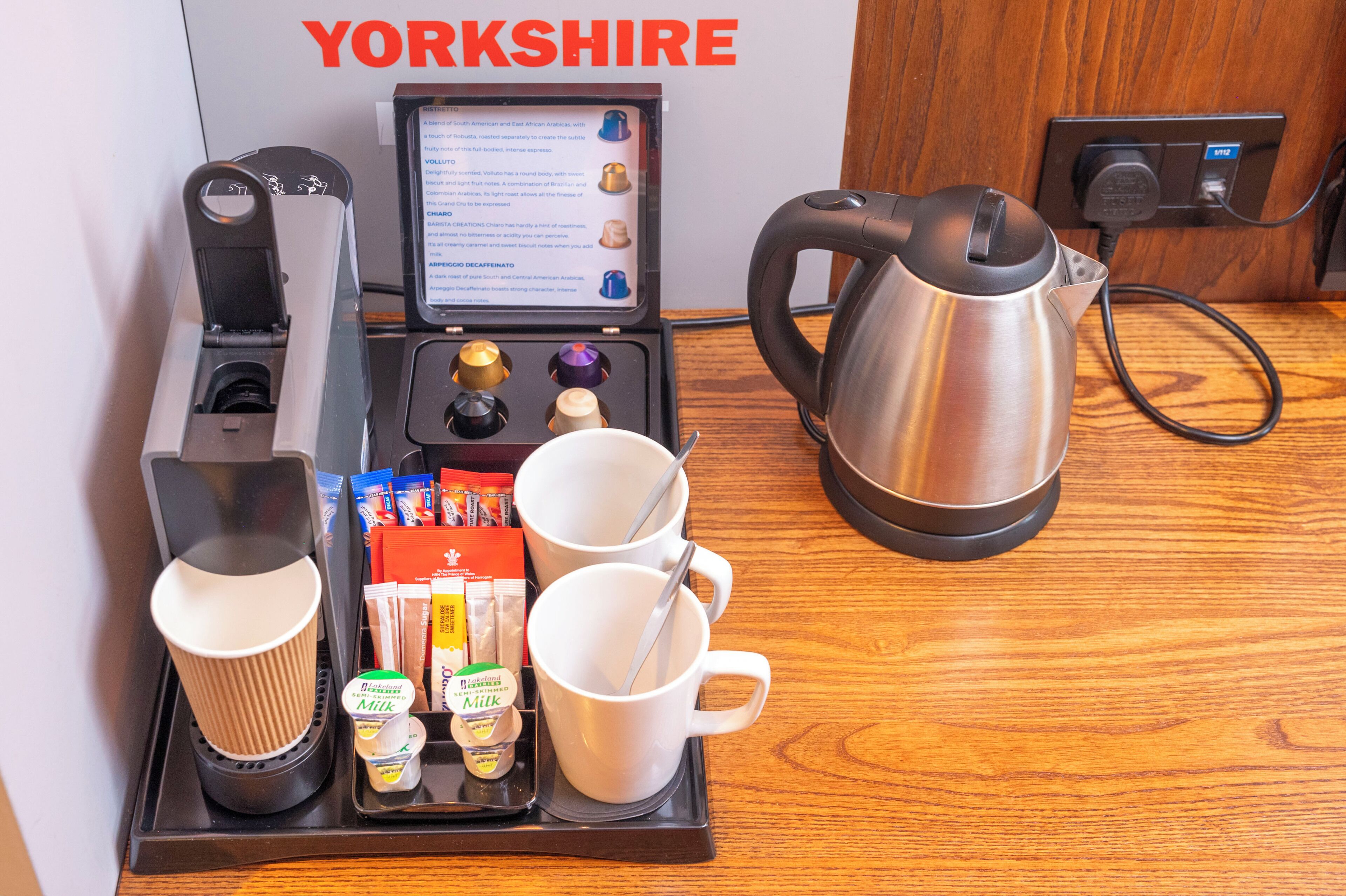 A breakfast setup with a coffee maker, tea maker, and a kettle on a tray, along with cups and milk containers.
