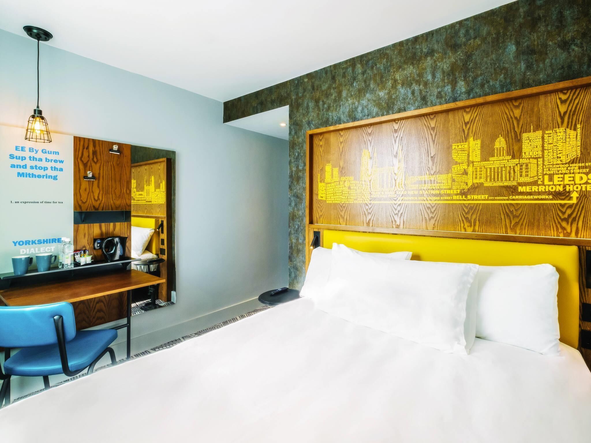 A hotel room with a bed, a desk with a chair, a coffee machine, and a wall with a cityscape design.