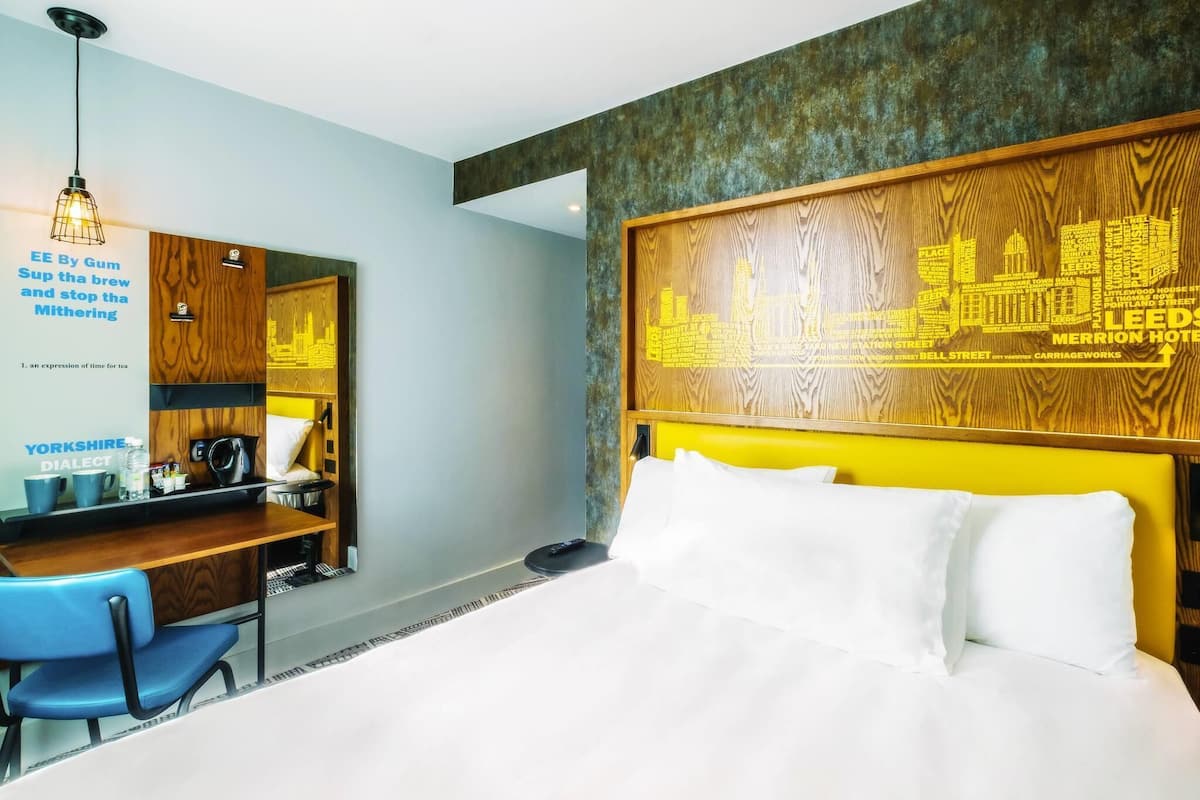 A hotel room with a bed, a desk with a chair, a coffee machine, and a wall with a cityscape design.
