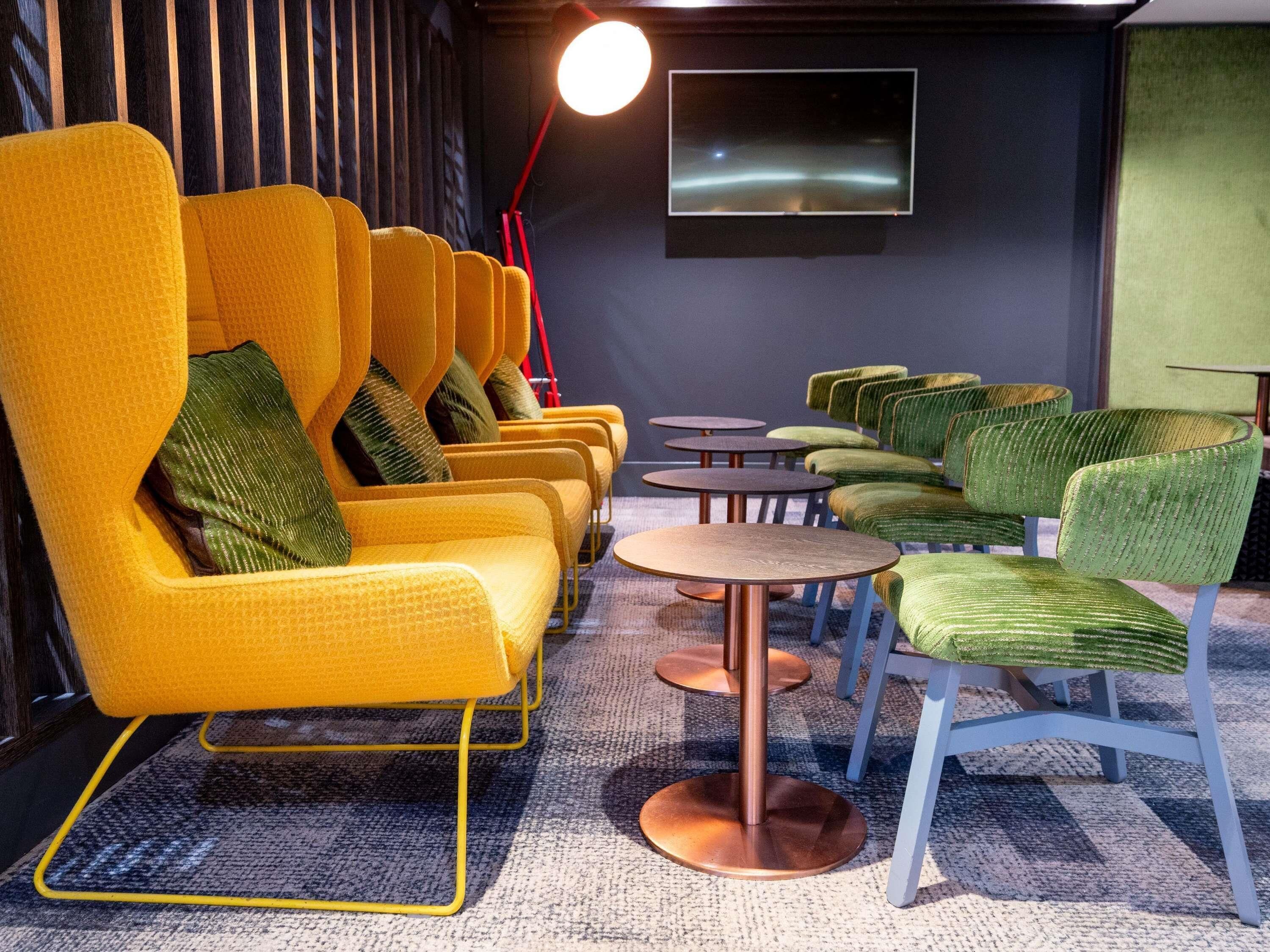 A modern lounge area with yellow and green chairs, small round tables, and a flat-screen TV on the wall.