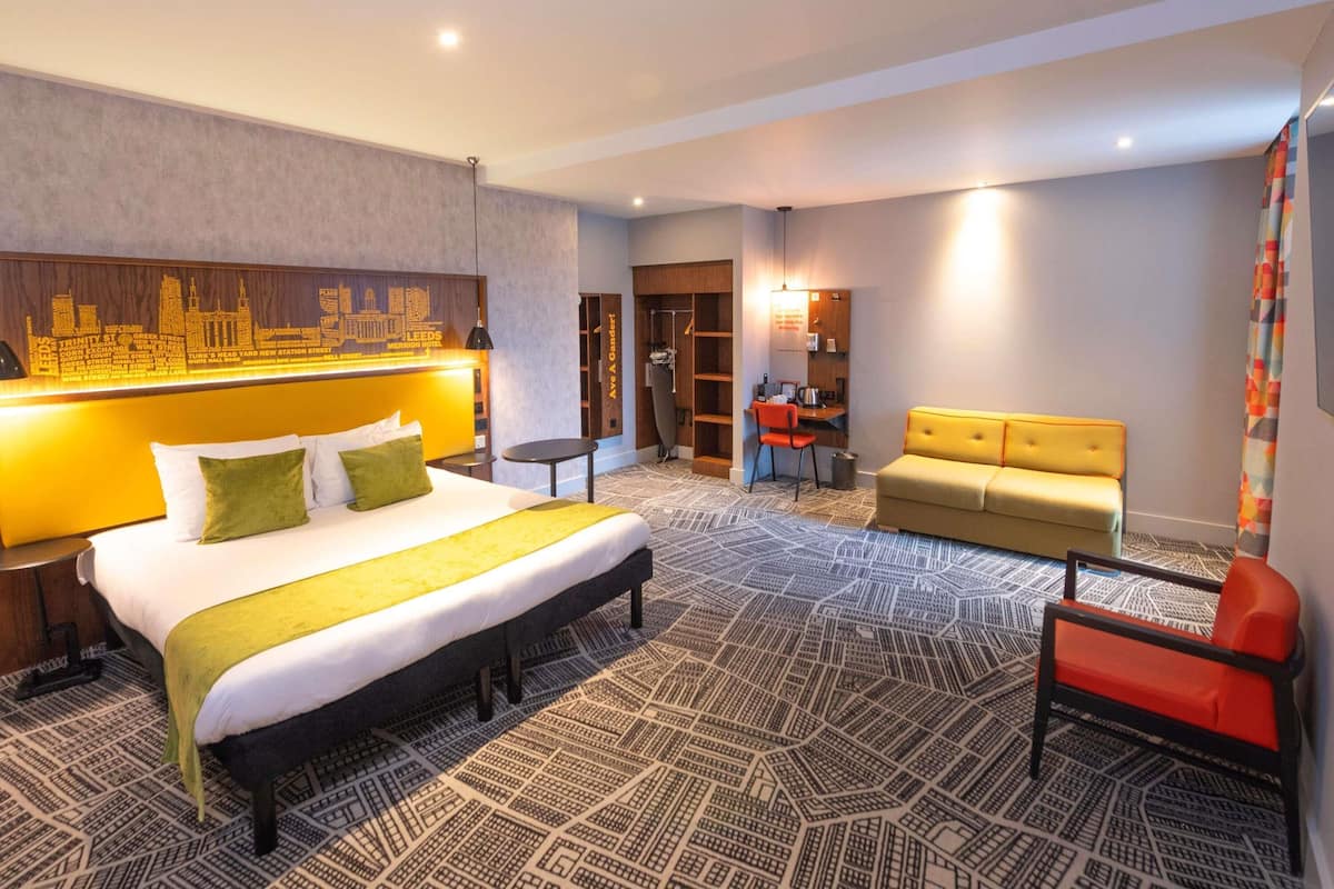 A hotel room with a large bed, a sofa, a chair, a desk, and a bookshelf.