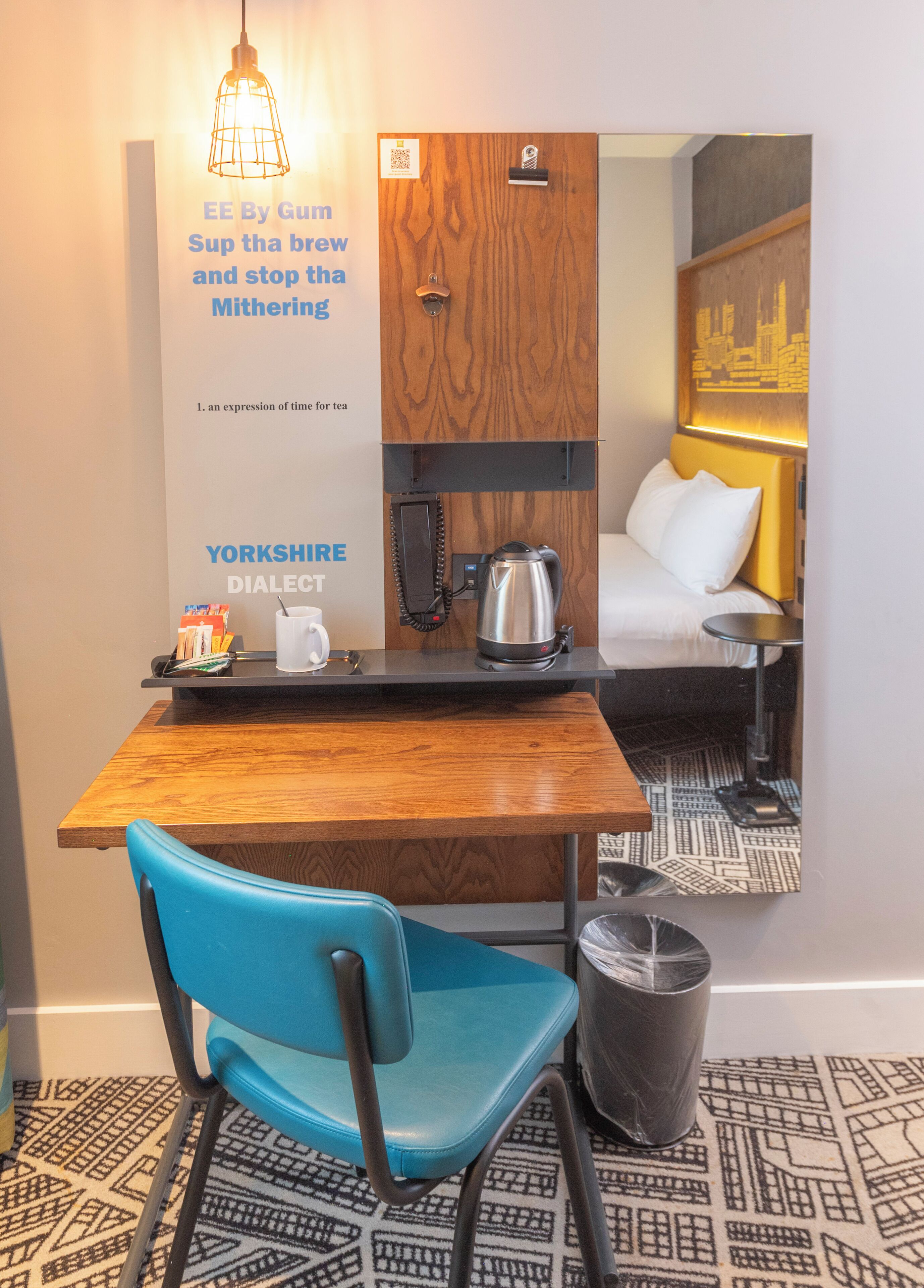 A hotel room with a wooden desk, a blue chair, a kettle, a phone, and a trash bin.