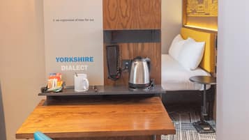 A hotel room with a wooden desk, a blue chair, a kettle, a phone, and a trash bin.