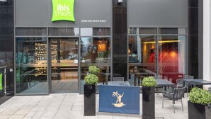 A modern building with a green 'ibis Styles' sign, glass doors, and outdoor seating area with tables and chairs.