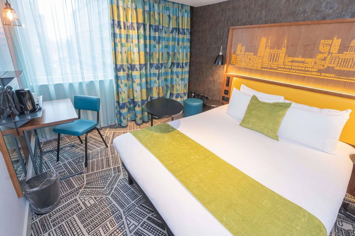A hotel room with a bed, a desk, a chair, a small table, and a trash bin.