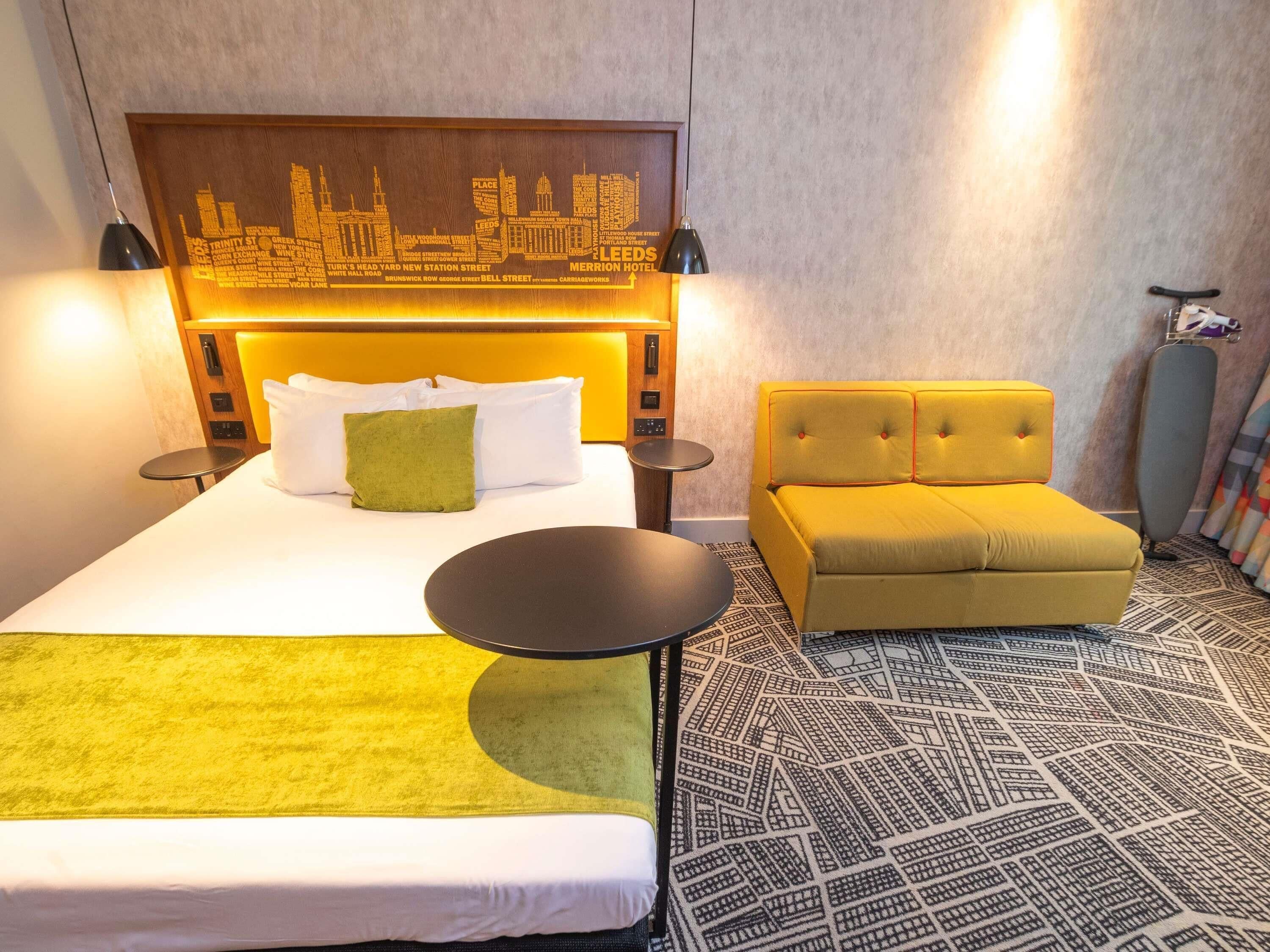 Ein Hotelzimmer mit einem Bett, einem gelben Sofa, einem runden Tisch und einem Stadtbild-Kunstwerk an der Kopfteil.
