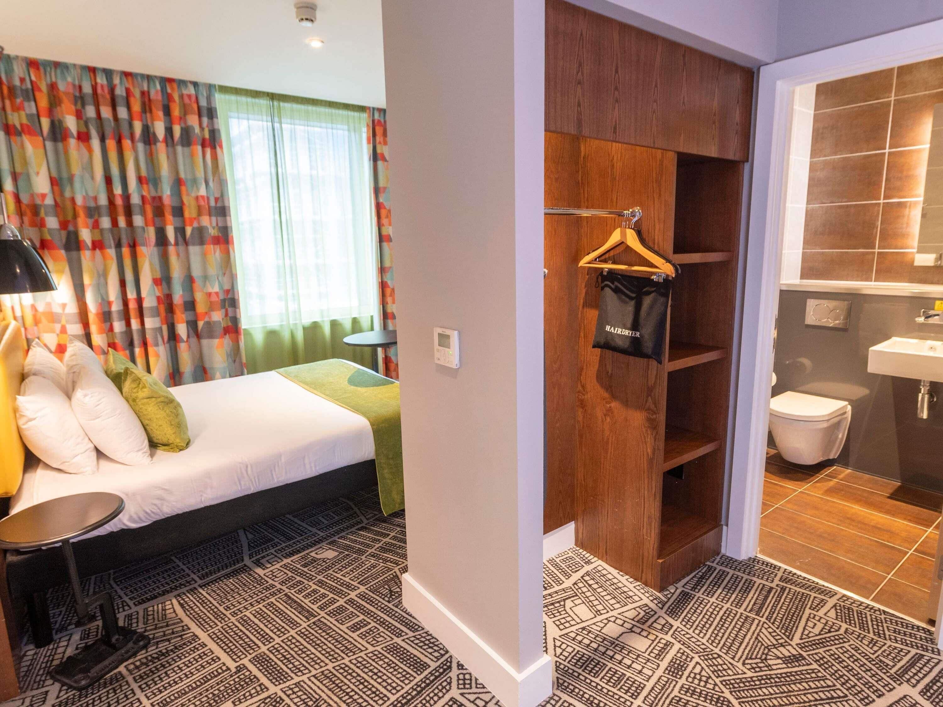 Une chambre d’hôtel comprenant un lit, une table de chevet, un sèche-cheveux mural et une salle de bain avec un lavabo et des toilettes.