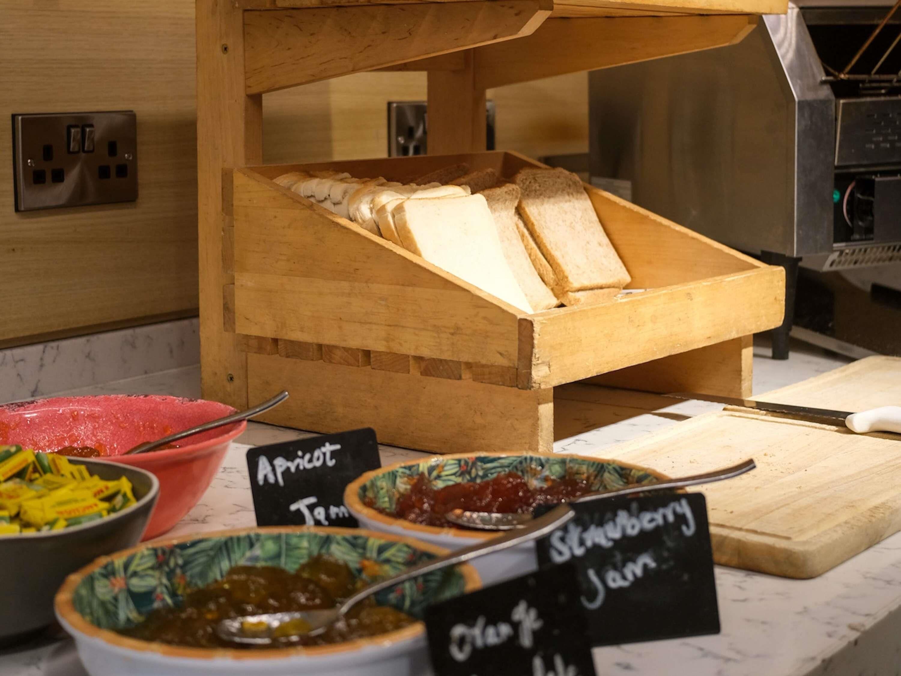 A wooden bread box with sliced bread, apricot jam, strawberry jam, and orange jelly on a countertop.