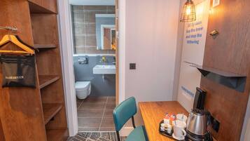 A compact hotel room with a wooden wardrobe, a small bathroom, a dining table with a coffee maker, and a blue chair.